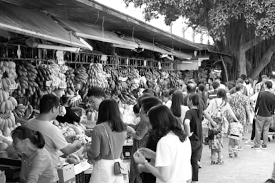 People participate in a bustling outdoor market with numerous stalls selling various fruits. The scene is busy with many individuals browsing and purchasing goods. Bunches of bananas and other tropical fruits hang prominently at the vendor stands.