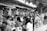 People participate in a bustling outdoor market with numerous stalls selling various fruits. The scene is busy with many individuals browsing and purchasing goods. Bunches of bananas and other tropical fruits hang prominently at the vendor stands.