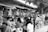 People participate in a bustling outdoor market with numerous stalls selling various fruits. The scene is busy with many individuals browsing and purchasing goods. Bunches of bananas and other tropical fruits hang prominently at the vendor stands.