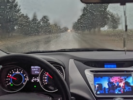 A car interior view with a dashboard displaying a digital speedometer, odometer, and fuel gauge. A screen on the dashboard shows a colorful image, possibly a video or media player. The windshield is covered with rain, providing a view of a road lined with trees under an overcast sky.
