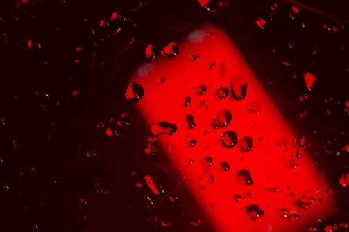 A vibrant red background is scattered with numerous droplets of liquid, creating an abstract and dynamic effect. The light and shadows enhance the contrast between the droplets and the deep red tones, providing a dramatic visual impact.