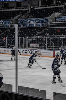 Hockey players in blue uniforms are on an indoor ice rink, actively engaged in a game. The venue appears to be a large, mostly empty stadium with tiered seating. Banners and advertisements, in both English and Cyrillic, line the walls and barriers around the rink. A few spectators are visible, and bright arena lights illuminate the scene.