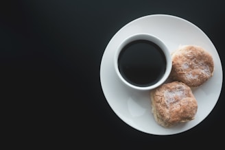 A white plate containing a cup of dark coffee and two biscuits is placed against a dark background. The biscuits have a flaky texture and appear freshly baked with a dusting of flour on top.