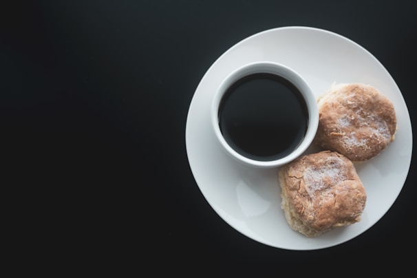 A white plate containing a cup of dark coffee and two biscuits is placed against a dark background. The biscuits have a flaky texture and appear freshly baked with a dusting of flour on top.