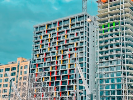 A set of modern high-rise buildings under construction with clear geometric patterns. The building in the center features a facade with vertical lines in red, yellow, and white, while the structure on the right is partially constructed, revealing its framework. The sky is a cloudy blue-green, creating a contrast with the buildings. There is minimal greenery visible at the base.