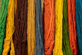Various strands of yarn are arranged vertically next to each other, showcasing an array of vibrant colors including green, brown, yellow, orange, blue, and more. Each strand's texture is visible with fine fibers, creating a visually rich tapestry.