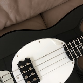 A close-up of a black and white electric bass guitar resting on a beige leather surface, showing the strings and a portion of the body.
