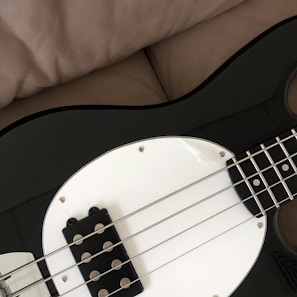 A close-up of a black and white electric bass guitar resting on a beige leather surface, showing the strings and a portion of the body.