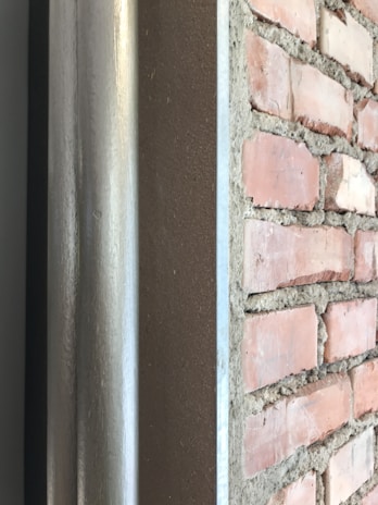 A detailed view of a brick wall with visible cement mortar between the bricks is depicted alongside a metal or concrete surface running parallel to it. The bricks have a reddish hue with a rough texture, while the mortar appears gray and uneven. The polished metal or concrete surface reflects some light, indicating its smooth finish.