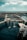 A waterfront scene featuring a dolphin enclosure with multiple dolphins mid-jump. Walkways and platforms provide access to the area. The background includes a rocky breakwater with an expansive ocean view and a partly cloudy sky.