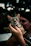 A person gently holding a tabby cat, with the background softly blurred. The cat's sharp gaze is directed toward the camera, while the person appears to be nuzzling close to the pet.