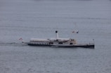 A vintage steamship is navigating through calm waters, displaying multiple flags, including an Austrian one at the stern. The vessel has a black hull and white superstructure with a tall smokestack at the center.