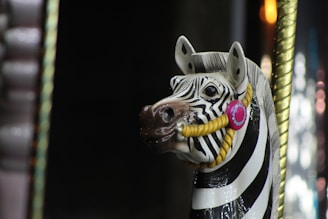 A carousel horse designed as a zebra with black and white stripes. It has a decorative bridle featuring a pink circular embellishment and a yellow rope. The background is dark with hints of gold and blurred lights.