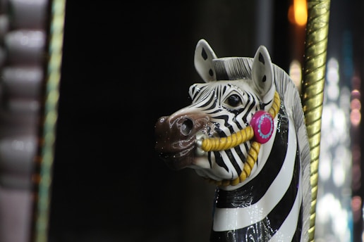A carousel horse designed as a zebra with black and white stripes. It has a decorative bridle featuring a pink circular embellishment and a yellow rope. The background is dark with hints of gold and blurred lights.