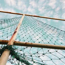 A net is stretched between rusty metal poles with a sky filled with clouds in the background. The net appears tangled at the bottom and is colored green.