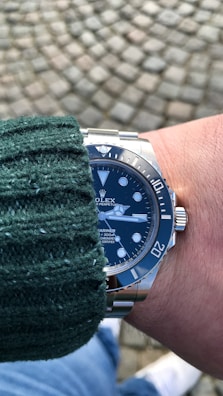 A wrist adorned with a luxury watch featuring a deep blue dial and a stainless steel band. The person is wearing a green knitted sweater, and the background consists of a cobblestone pavement.