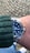 A wrist adorned with a luxury watch featuring a deep blue dial and a stainless steel band. The person is wearing a green knitted sweater, and the background consists of a cobblestone pavement.