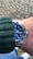 A wrist adorned with a luxury watch featuring a deep blue dial and a stainless steel band. The person is wearing a green knitted sweater, and the background consists of a cobblestone pavement.