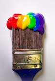 A paintbrush with thick, vibrant blobs of paint in red, orange, yellow, green, blue, and purple covering the bristles, against a plain background.