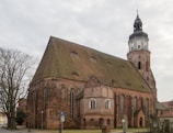 A large, historical brick church with a tall, pointed roof and a prominent bell tower. The architecture features Gothic-style windows and intricate brickwork. The setting includes a bare tree to the left and a parking sign in front.