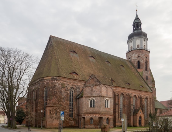 A large, historical brick church with a tall, pointed roof and a prominent bell tower. The architecture features Gothic-style windows and intricate brickwork. The setting includes a bare tree to the left and a parking sign in front.