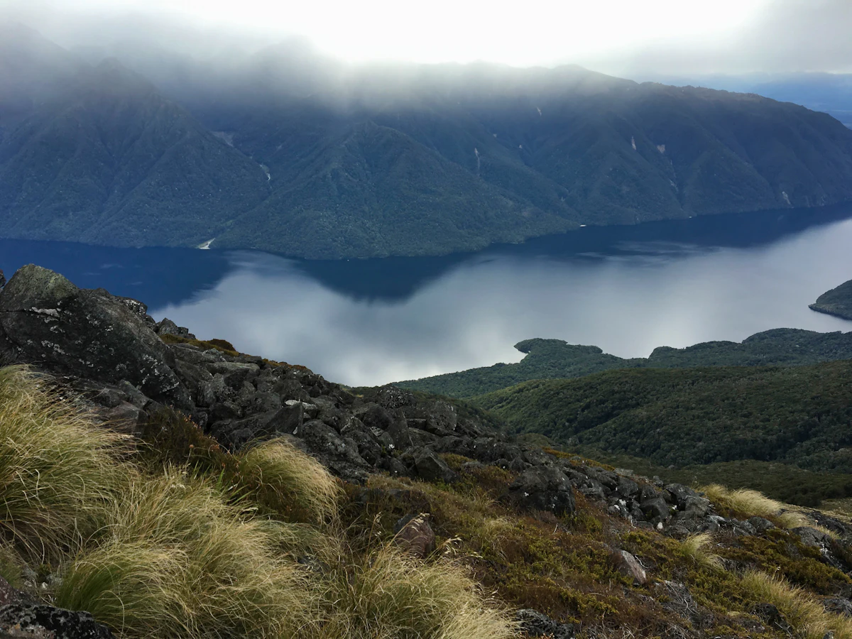 A lake far below a rocky, tussock-covered slope near Te Anau, with cloud on the forested hills across the water