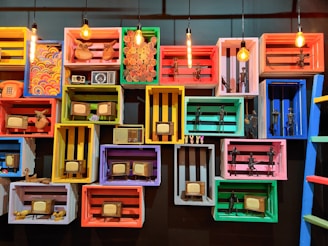 A collection of colorful wooden crates mounted on a wall, each containing vintage items such as retro televisions, radios, and small figurines. The crates are painted in bright colors including yellow, red, blue, green, and pink, with some containing decorative patterns. Several light bulbs hang above, illuminating the display.