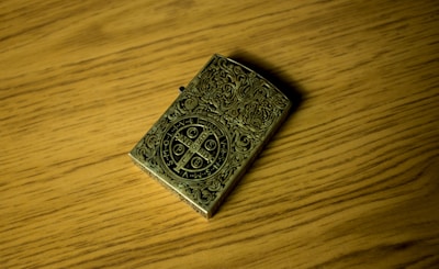 An intricately designed metallic lighter rests on a wooden surface. The lighter features elaborate engravings and a central circular emblem with various symbols and letters.
