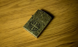 An intricately designed metallic lighter rests on a wooden surface. The lighter features elaborate engravings and a central circular emblem with various symbols and letters.