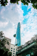 A tall modern skyscraper with a unique helipad design ascends into a partly cloudy blue sky. Surrounding the building are lush green leaves, framing the top of the image. At the bottom, there are signs for 'Saigon Garden' and 'Saigon Prince Hotel' indicating the location.