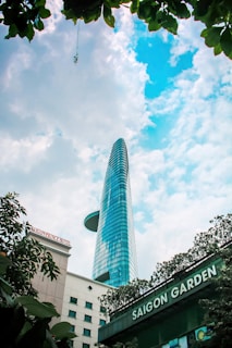 A tall modern skyscraper with a unique helipad design ascends into a partly cloudy blue sky. Surrounding the building are lush green leaves, framing the top of the image. At the bottom, there are signs for 'Saigon Garden' and 'Saigon Prince Hotel' indicating the location.