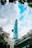 A tall modern skyscraper with a unique helipad design ascends into a partly cloudy blue sky. Surrounding the building are lush green leaves, framing the top of the image. At the bottom, there are signs for 'Saigon Garden' and 'Saigon Prince Hotel' indicating the location.