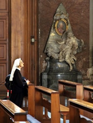 A nun is kneeling in prayer in front of a beautifully carved religious monument inside a church. The monument features a sculpted figure, possibly an angel, and a portrait set within an ornate frame. Sunlight streams through, illuminating the scene with a serene glow. The wooden pews and rich cathedral architecture add to the sense of reverence and tranquility.