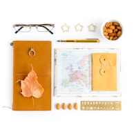 An arrangement of various objects on a white background includes a leather-bound notebook with a leaf and a ring on top, reading glasses, a small map of Europe, a sealed envelope, a pen, a ceramic bowl of almonds, and star-shaped paper clips. There is also a stencil with numbers and symbols and several almonds laid out in a row.