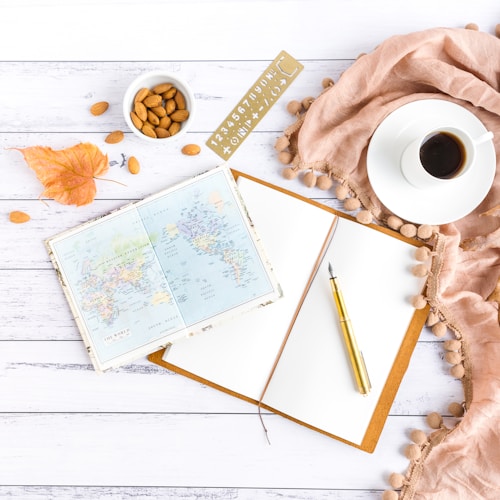An open blank notebook lies on a white wooden surface, accompanied by a gold fountain pen. To its left, a world map is spread open, adding a sense of travel and exploration. Near the map, a small white bowl is filled with almonds, with some scattered around it. A dry autumn leaf adds a hint of seasonal change. A cup of black coffee sits on a saucer, beside a tassel-edged beige fabric, providing a cozy, warm feel. A stencil with letters and numbers completes the scene, suggesting creative planning or journaling.