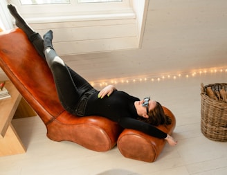 A person is lounging on a brown leather chaise longue with a relaxed pose, wearing black clothing and sunglasses. The setting is indoors with a cozy atmosphere, illuminated by warm string lights along the wall. A wicker basket filled with logs is positioned nearby, and sunlight filters through a window above.