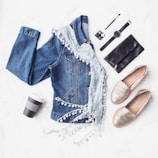 Flat lay of fenr clothing items including a premium jacket and custom jeans on a simple, muted surface.