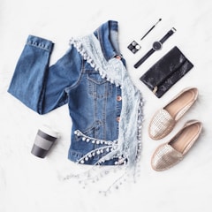 A flat lay of fashion items including a blue denim jacket adorned with a light lace scarf, a pair of beige woven loafers, a black wallet, a black wristwatch, eye shadow, an eyeliner, and a take-away coffee cup placed on a fluffy white surface.