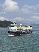 A passenger ferry is sailing on clear blue waters with a backdrop of a mountainous coastline under a partly cloudy sky. The ferry is painted in blue and white, with black tires attached to its sides and a red lower hull.