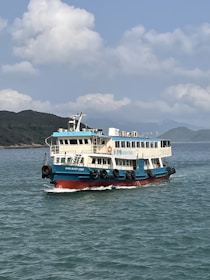 A passenger ferry is sailing on clear blue waters with a backdrop of a mountainous coastline under a partly cloudy sky. The ferry is painted in blue and white, with black tires attached to its sides and a red lower hull.