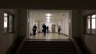 A dimly lit hallway with several people walking and standing. The setting appears to be a hospital or institutional building with white walls and a tiled ceiling. A few individuals are visible, some wearing masks, suggesting a precautionary measure. The floor is spacious, and the lighting overhead provides a soft glow.