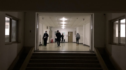 A dimly lit hallway with several people walking and standing. The setting appears to be a hospital or institutional building with white walls and a tiled ceiling. A few individuals are visible, some wearing masks, suggesting a precautionary measure. The floor is spacious, and the lighting overhead provides a soft glow.