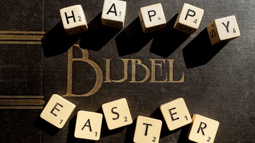 Wooden Scrabble tiles spell out 'Happy Easter' arranged over a dark surface with the word 'BIBLE' visible in gold lettering underneath. The tiles cast soft shadows, creating a warm and inviting atmosphere.