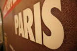 A close-up of a textured surface features the word 'PARIS' in large white letters. The background appears to be a mosaic pattern with shades of brown, red, and hints of other colors.
