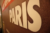 A close-up of a textured surface features the word 'PARIS' in large white letters. The background appears to be a mosaic pattern with shades of brown, red, and hints of other colors.