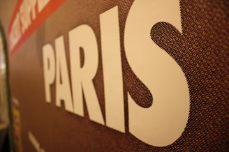 A close-up of a textured surface features the word 'PARIS' in large white letters. The background appears to be a mosaic pattern with shades of brown, red, and hints of other colors.