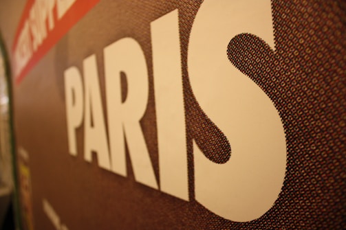 A close-up of a textured surface features the word 'PARIS' in large white letters. The background appears to be a mosaic pattern with shades of brown, red, and hints of other colors.