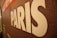 A close-up of a textured surface features the word 'PARIS' in large white letters. The background appears to be a mosaic pattern with shades of brown, red, and hints of other colors.