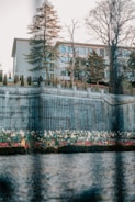 A multi-story building is surrounded by tall trees and a manicured garden. A stone wall with metal railings runs along the edge of the garden, and colorful flowers are visible in the foreground. The scene is reflected in a body of water, adding to the serene atmosphere.