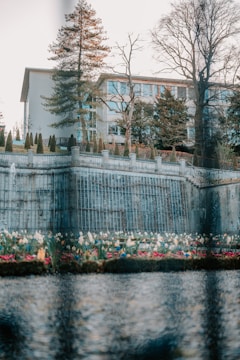 A multi-story building is surrounded by tall trees and a manicured garden. A stone wall with metal railings runs along the edge of the garden, and colorful flowers are visible in the foreground. The scene is reflected in a body of water, adding to the serene atmosphere.